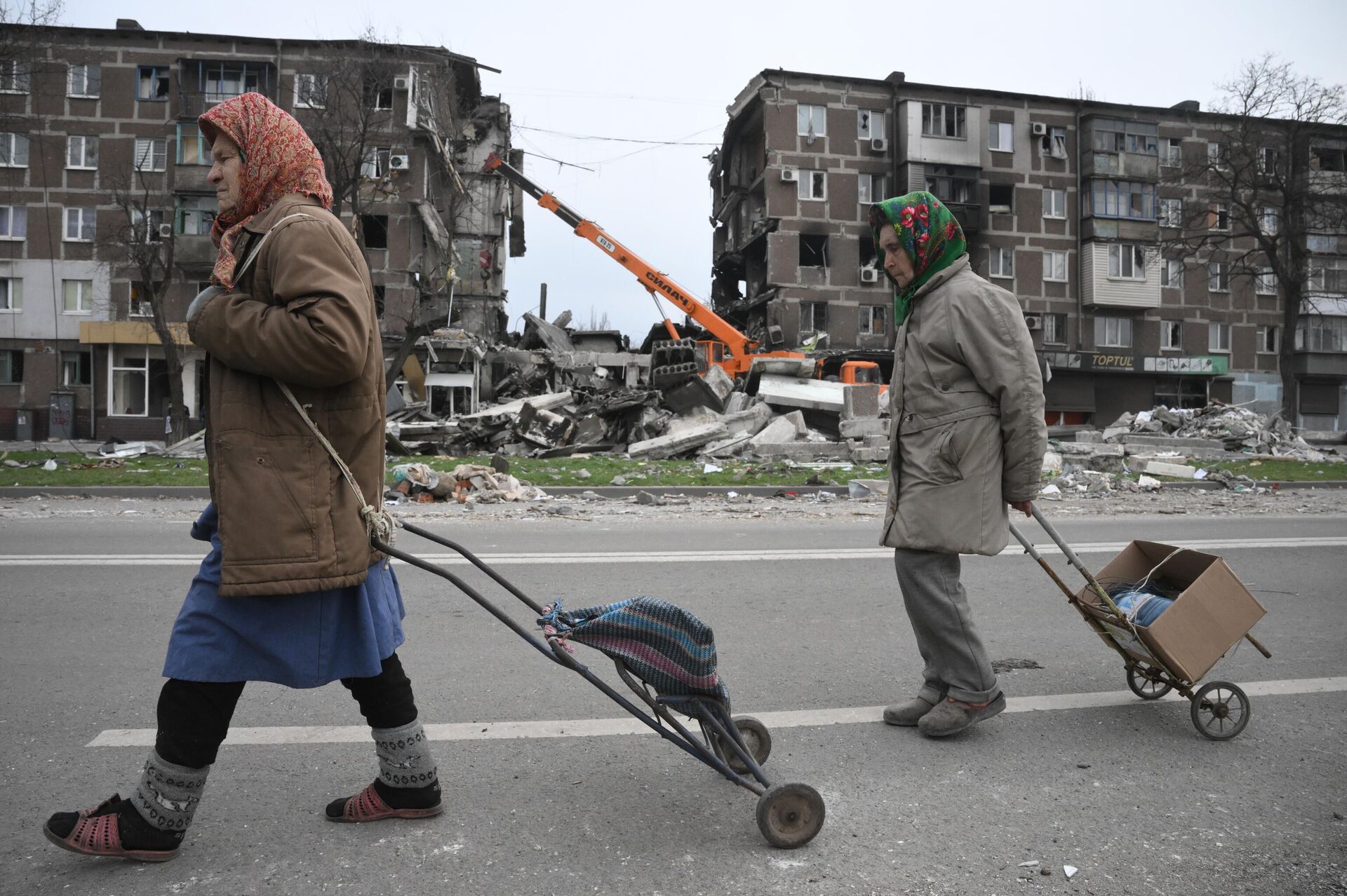Жители Мариуполя идут по улице города. На втором плане: сотрудники МЧС ДНР разбирают завалы у разрушенных домов - Sputnik Узбекистан, 1920, 13.04.2022