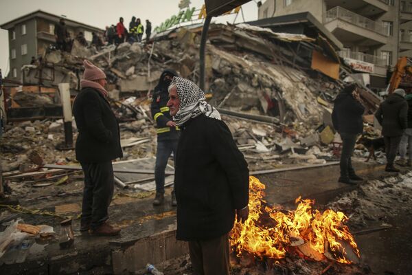 Люди греются рядом с рухнувшим зданием в Малатье, Турция, вторник, 7 февраля 2023 года. - Sputnik Узбекистан