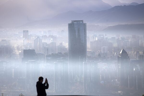 Шапка смога над крупными городами хорошо просматривается с возвышенностей недалеко от мегаполисов. Мужчина фотографирует облако смога над Сантьяго, Чили, июль 2018 года.  - Sputnik Узбекистан