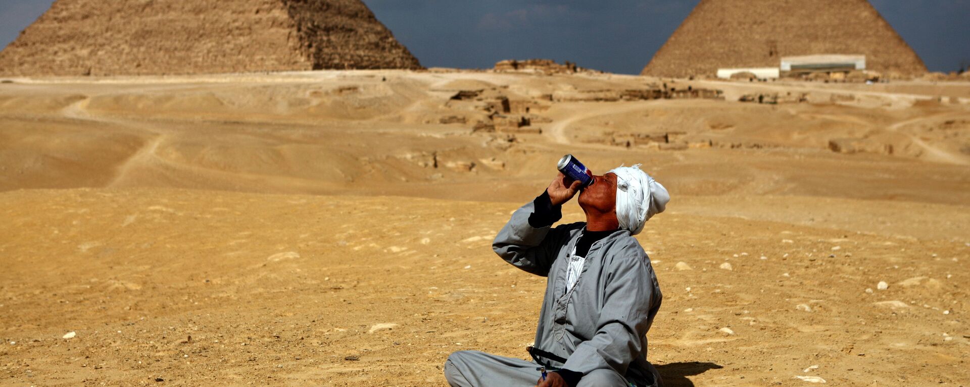 Мужчина пьет газированный напиток у пирамид в Гизе - Sputnik Узбекистан, 1920, 24.06.2024