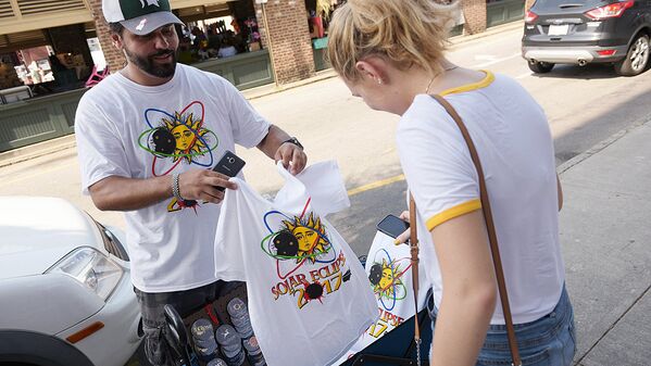T-shirt vendor Jan Dahouas shows an eclipse t-shirt of his own design to a buyer on a street near the City Market in Charleston, South Carolina, on August 20, 2017. - Sputnik Узбекистан